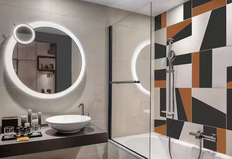Badezimmer mit beleuchtetem Rundspiegel, Waschtisch und Dusche mit geometrischen Fliesen.