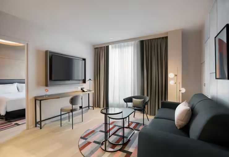 Sitzecke im Hotelzimmer mit dunklem Sofa, Sessel, zwei kleinen Tischen und Schreibtisch mit Fernseher.