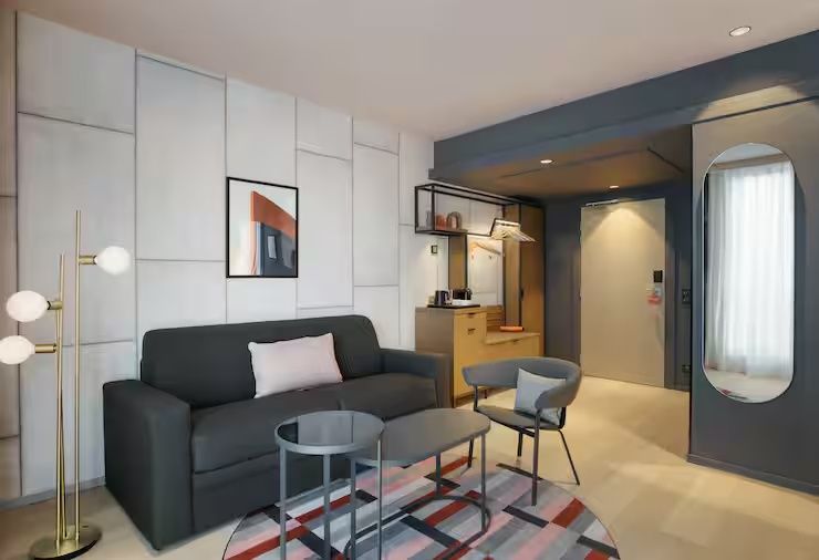 Wohnbereich mit dunklem Sofa, grauem Sessel, runden Tischen auf einem Teppich sowie Garderobe im Eingang.