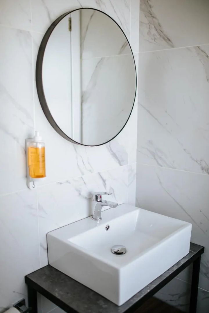 Weißes Waschbecken mit Armatur unter einem runden Spiegel an einer Wand in Marmoroptik.