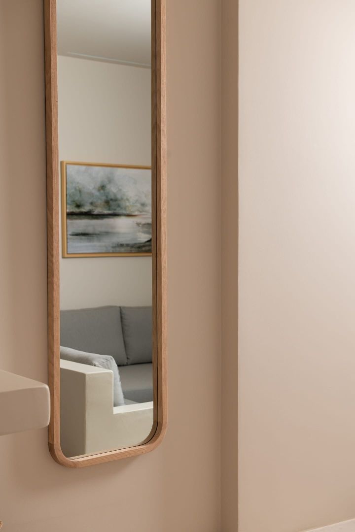 Ein schmaler Wandspiegel mit Holzrahmen reflektiert ein graues Sofa und ein Bild an einer beigen Wand.