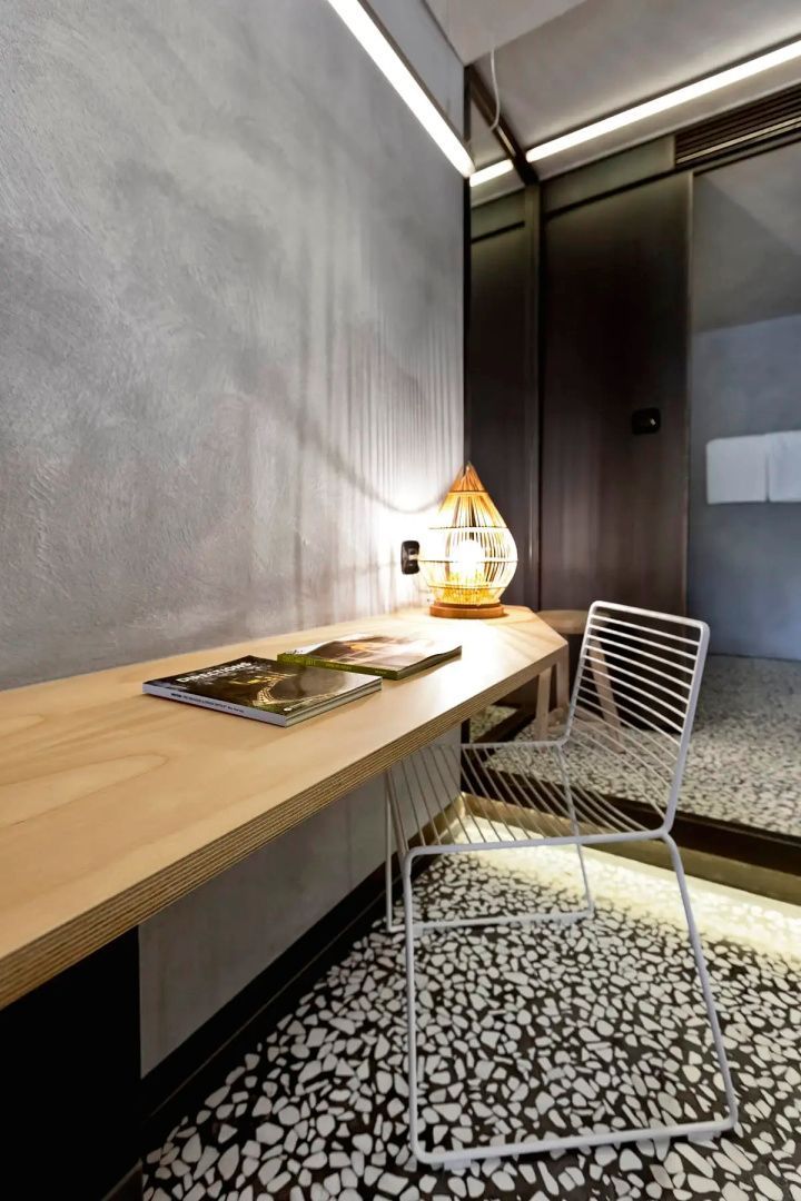 Schreibtisch aus Holz mit weißem Metallstuhl, Tischleuchte und grauer Wand im Zimmer.