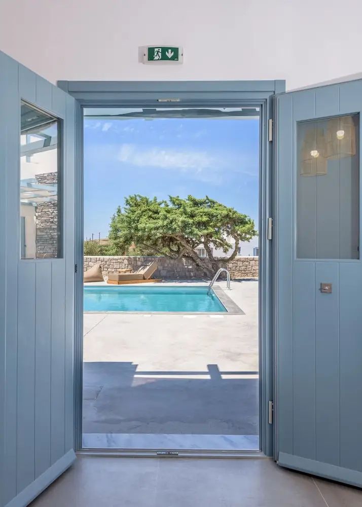 Blick durch die geöffnete blaue Tür auf eine Terrasse mit Swimmingpool und Liegen.
