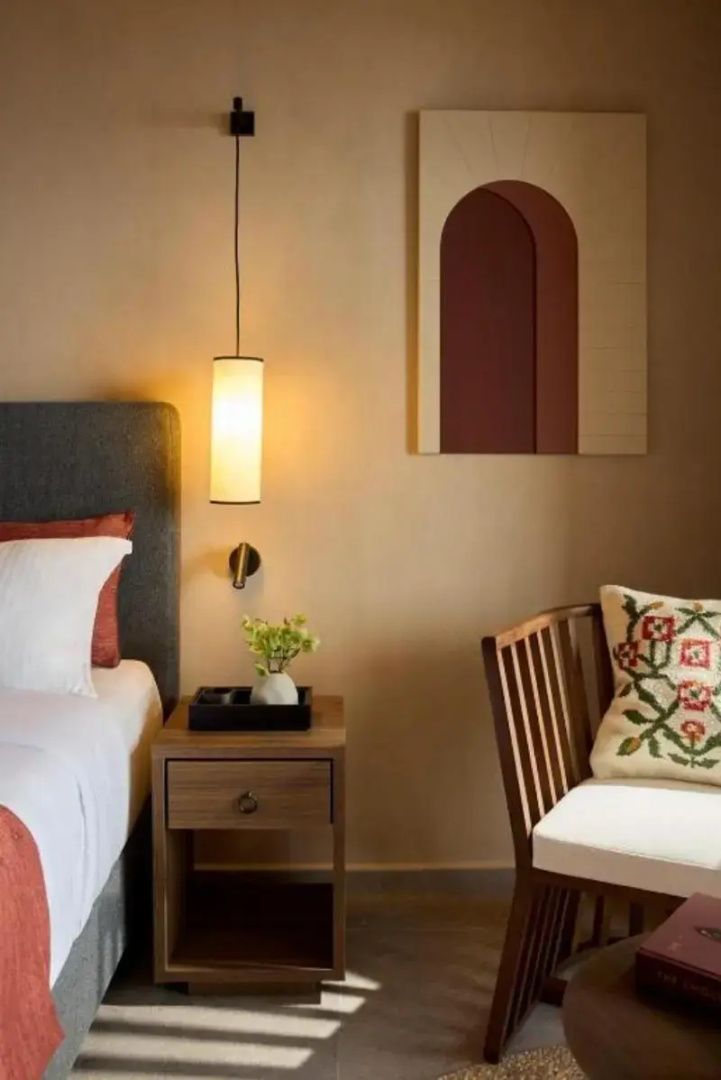Hotelzimmer mit Bett, Holznachttisch, Hängelampe und verziertem Kissen auf dem Stuhl.