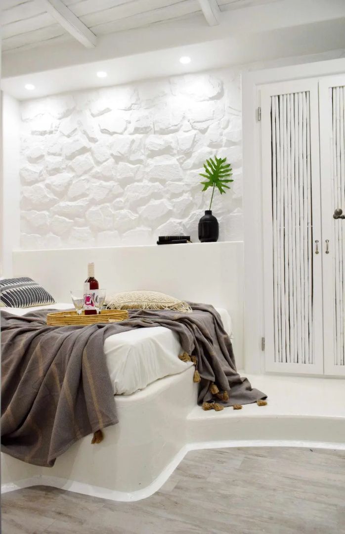 Weiß gestaltetes Hotelzimmer mit Doppelbett, Natursteinwand und Holztablett mit Weinflasche.