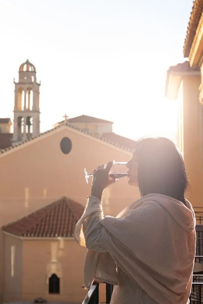 Eine Frau sitzt auf dem Balkon und trinkt ein Glas Wein mit Blick auf einen Kirchturm.