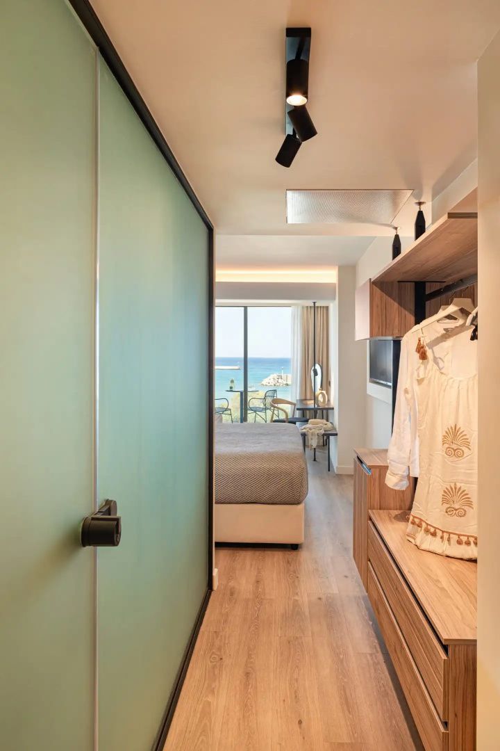 Blick durch ein modernes Hotelzimmer mit Garderobe und Bett in Richtung Balkon mit Meerblick.