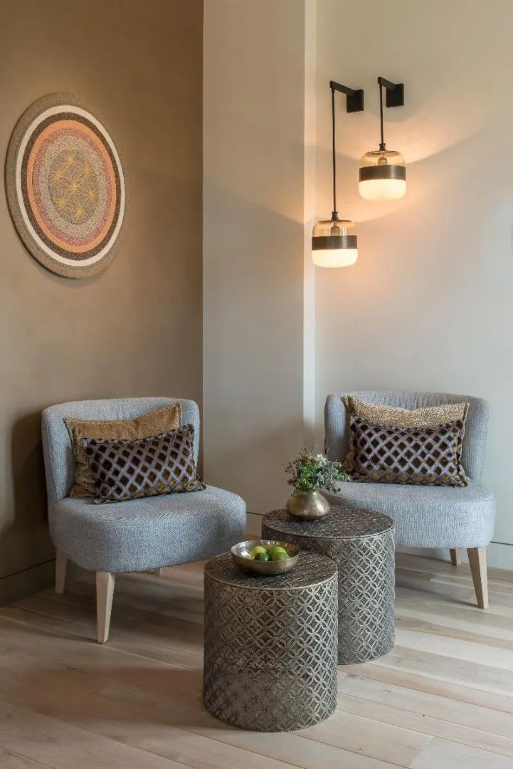 Zwei graue Sessel mit gemusterten Kissen und metallene Beistelltische in einer Ecke des Raumes.