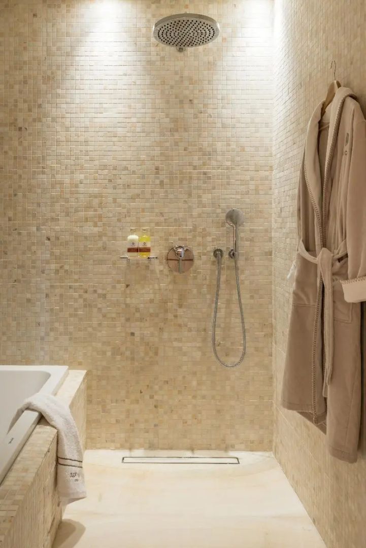 Dusche mit beigefarbenen Mosaikfliesen, Duscharmatur und einem aufgehängten Bademantel.