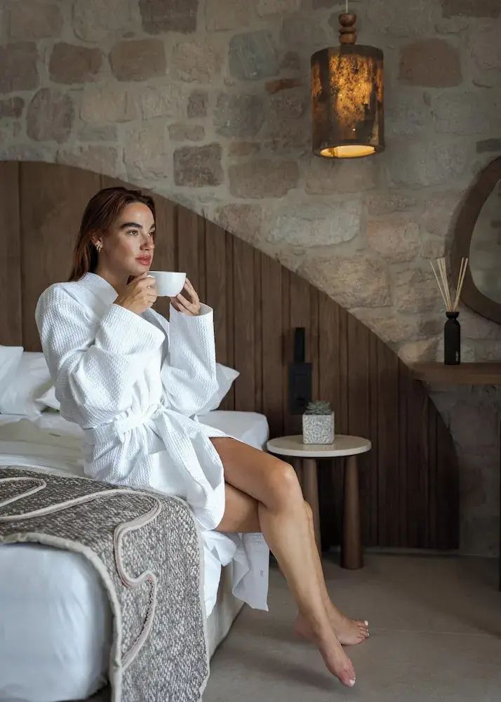 Eine Frau im weißen Bademantel sitzt auf dem Bett und trinkt aus einer Kaffeetasse.