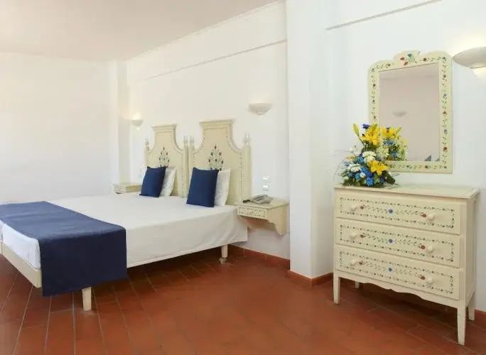 Helles Hotelzimmer mit Doppelbett, blauer Tagesdecke, Fliesenboden und verzierter Kommode mit Blumenstrauß.