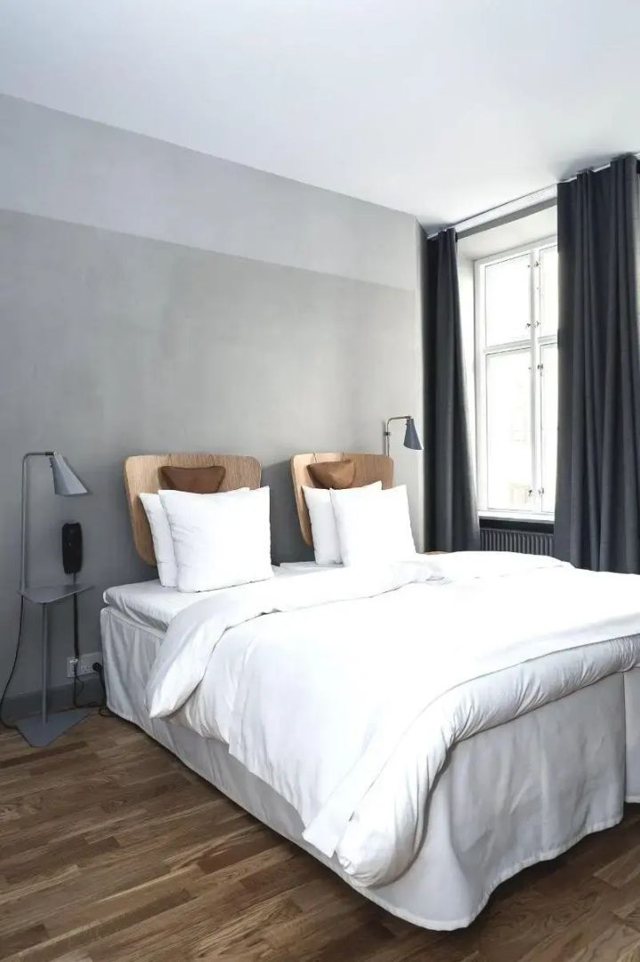 Doppelbett mit weißer Bettwäsche und Holz-Kopfteilen an einer grauen Wand neben einem Fenster.
