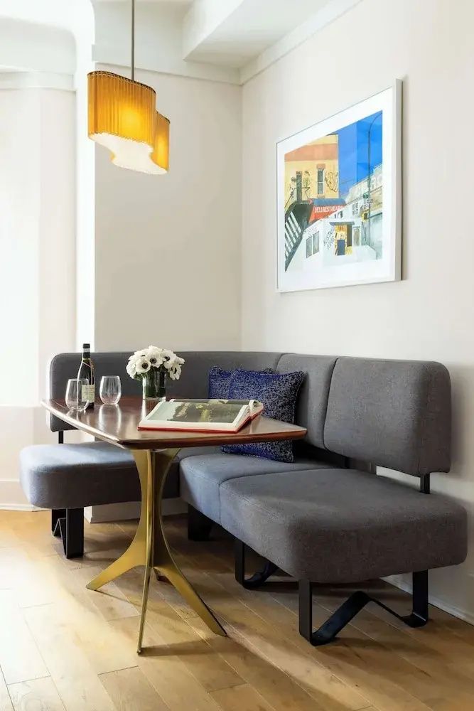 Sitzecke im Zimmer mit grauer Polstereckbank, Holztisch und buntem Bild an der Wand.