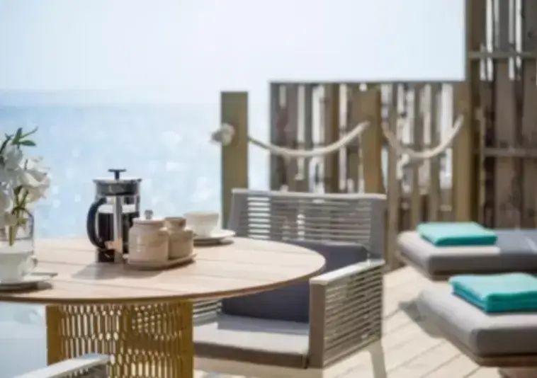 Sonnige Holzterrasse mit Tisch, Stuhl und Kaffeekanne direkt am türkisfarbenen Meer.