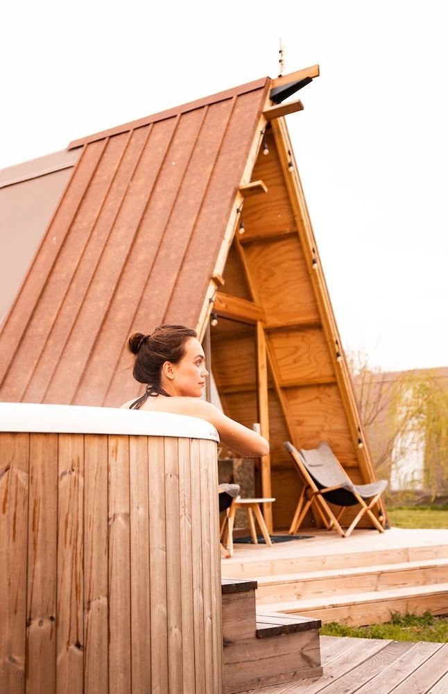 Frau sitzt in einem hölzernen Whirlpool vor einer A-Frame-Unterkunft aus Holz.