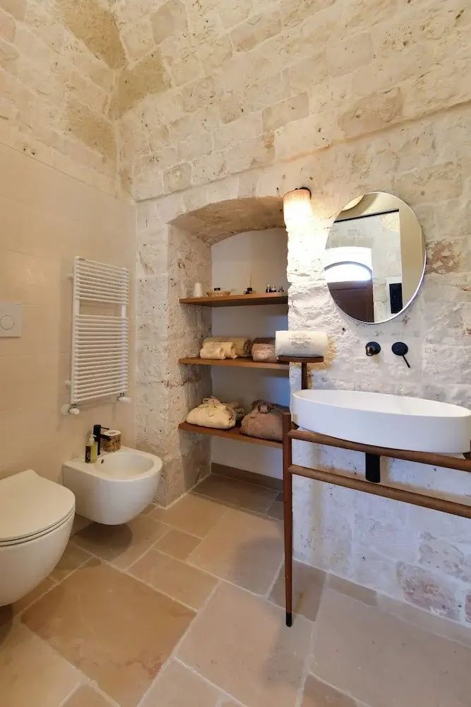 Badezimmer mit hellen Natursteinwänden, ovalem Waschtisch, Toilette und Bidet.