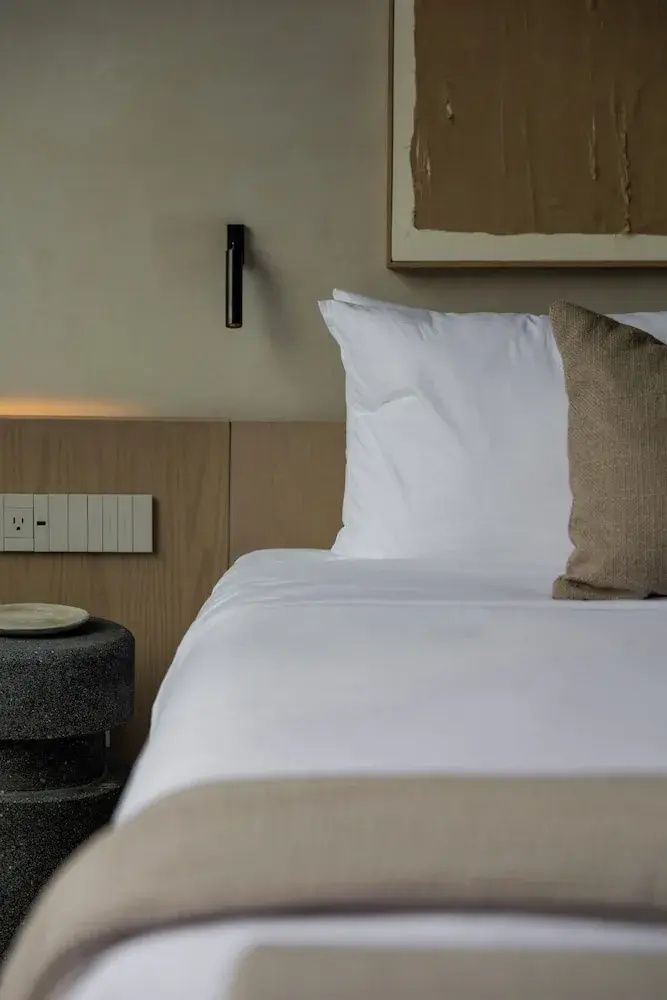 Doppelbett mit weißer Bettwäsche, beigefarbenem Kissen und Holzanschlussleiste im Zimmer.