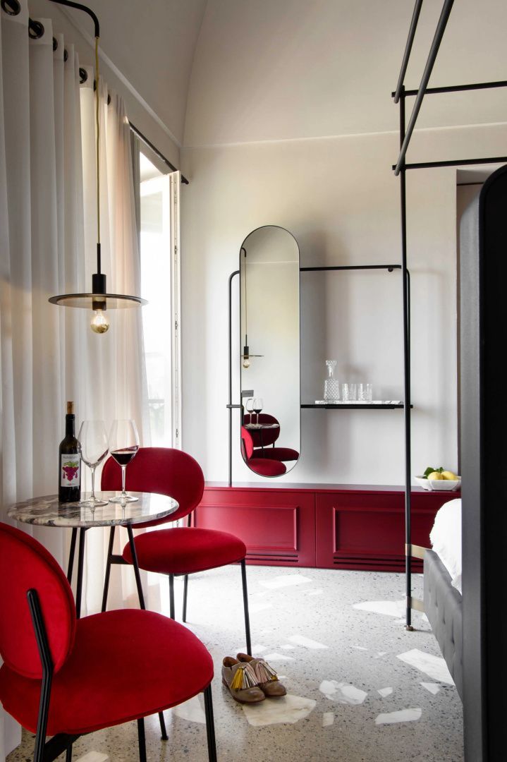 Sitzbereich im Hotelzimmer mit roten Stühlen, Marmortisch, Weingläsern und ovalem Wandspiegel.