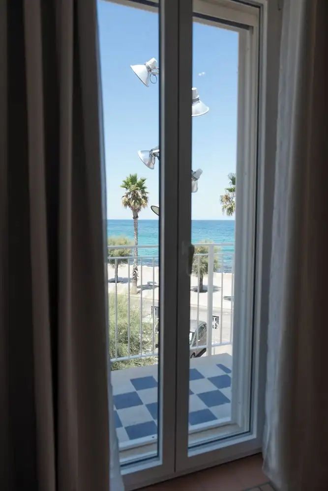 Blick durch eine Balkontür auf das blaue Meer, Palmen und eine Küstenpromenade.