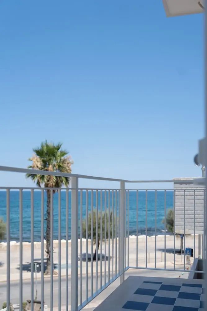 Blick von einem Balkon mit weißem Geländer auf das blaue Meer und Palmen unter blauem Himmel.