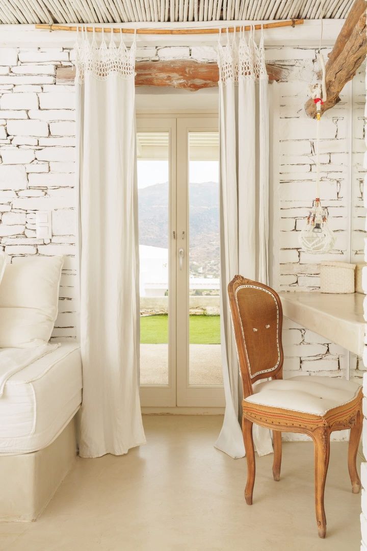 Heller Hotelraum mit weißer Steinwand, Holzstuhl am Schreibtisch und Vorhängen vor der Balkontür.