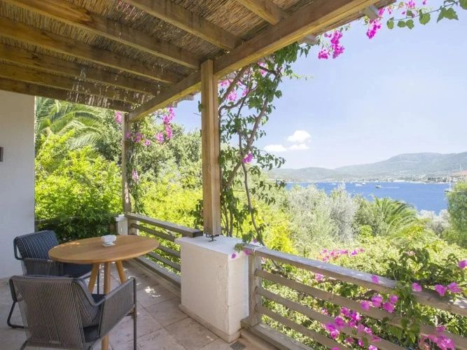 Überdachte Terrasse mit Pergola, Sitzgelegenheit, rosa Blüten und Ausblick auf das Meer.