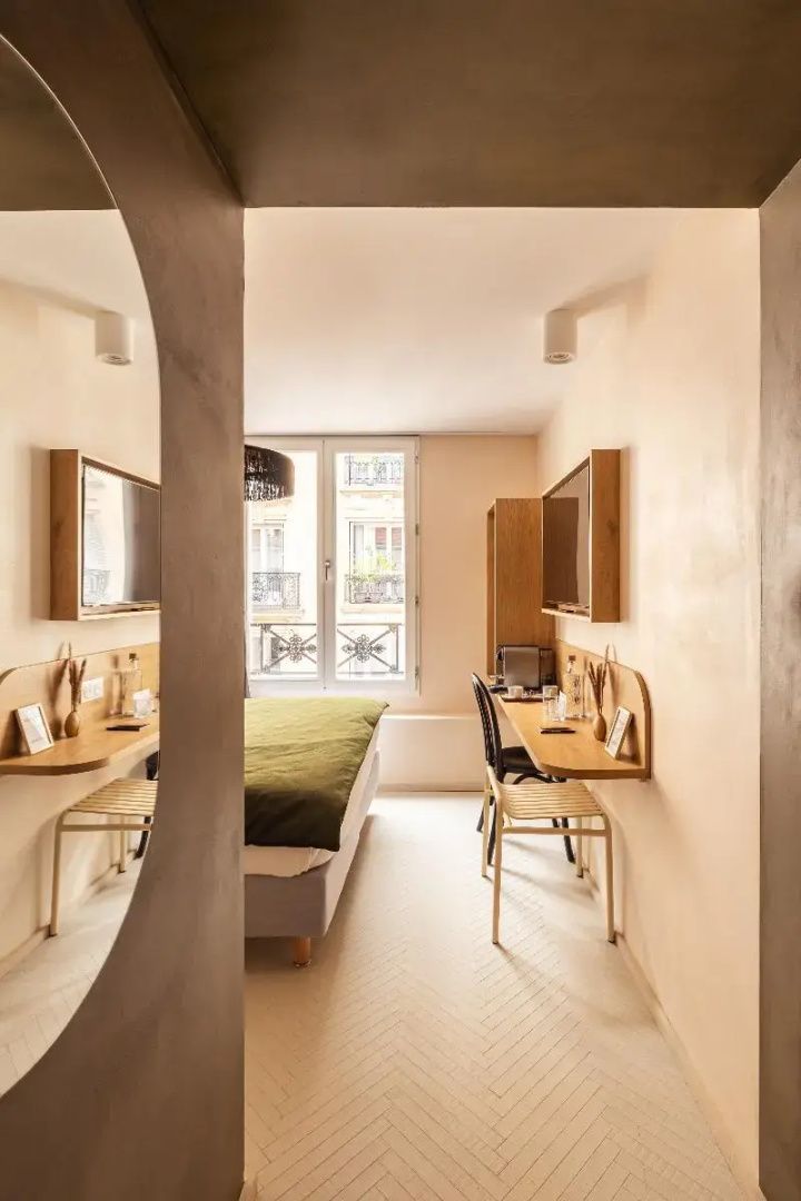 Helles Hotelzimmer mit Doppelbett, Wandtisch und Blick auf das gegenüberliegende Gebäude.