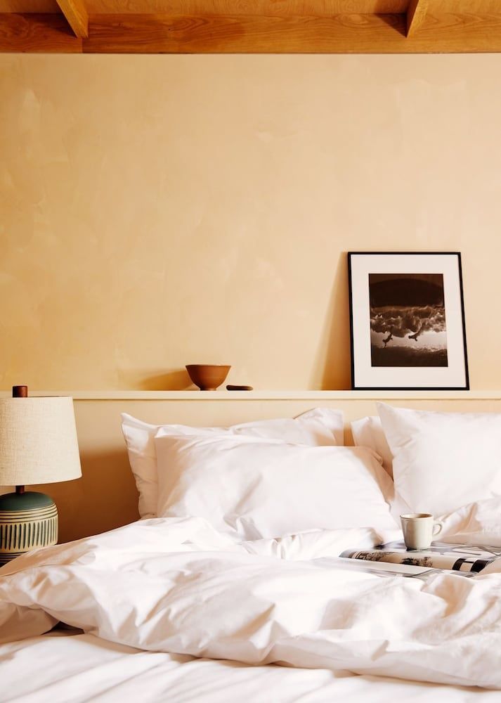 Bett mit weißer Bettwäsche, Nachttischlampe und eingerahmtem Bild an einer beigen Wand.