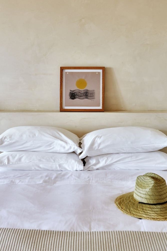 Doppelbett mit weißen Kissen, Strohhut und Wandbild mit Sonnenmotiv.