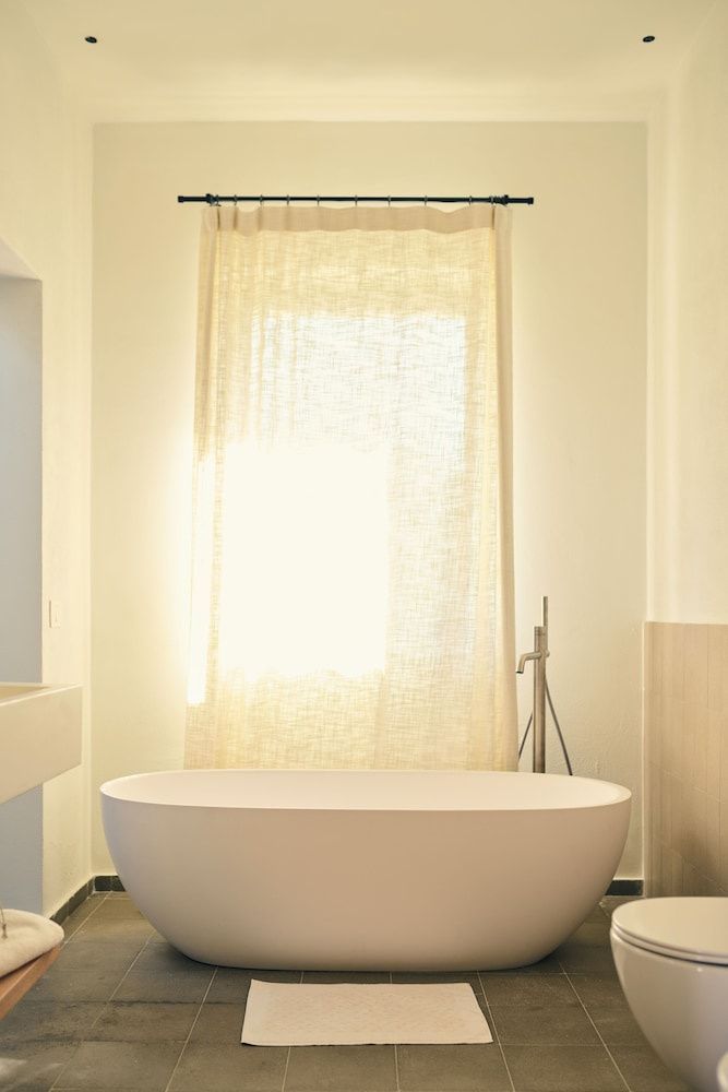 Freistehende weiße Badewanne vor einem hellerleuchteten Fenster mit Stoffvorhang.