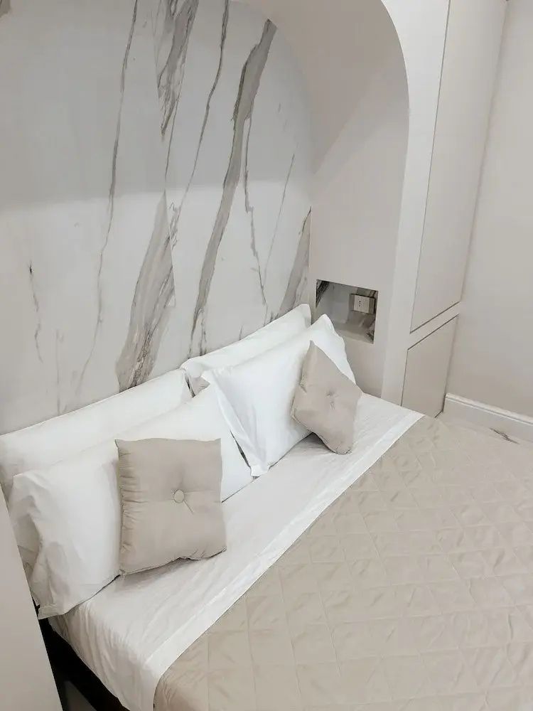 Bett mit weißen Kissen und beigen Dekokissen vor einer Wand in Marmoroptik.