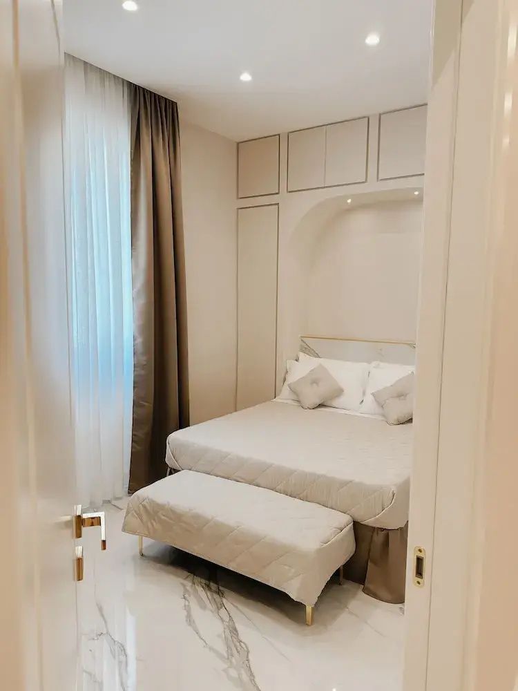 Doppelbett mit hellbrauner Tagesdecke, einer gepolsterten Bank davor und hellen Vorhängen am Fenster.