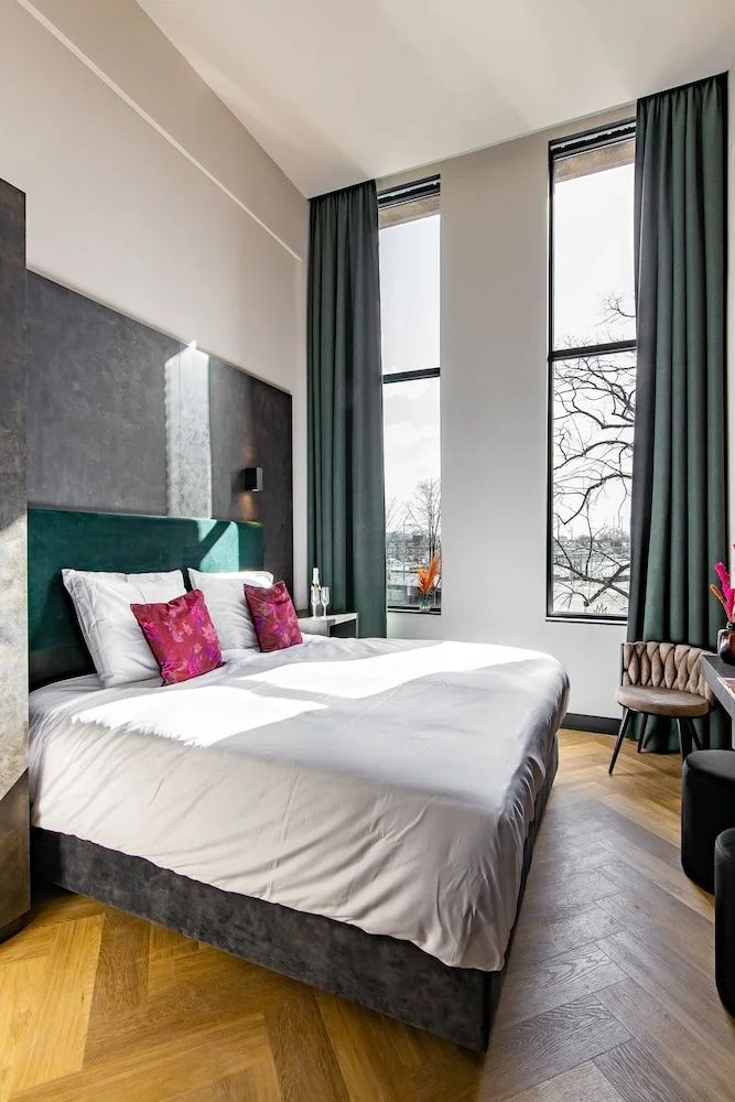 Helles Hotelzimmer mit großem Doppelbett, hohen Fensterfronten und grünen Vorhängen.