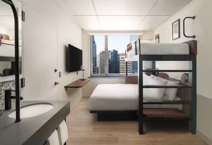 Modernes Hotelzimmer mit Doppelbett, Etagenbett und Fenster mit Blick auf Hochhäuser.
