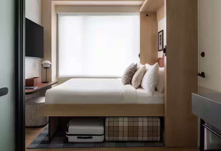Doppelbett in einem hellen Hotelzimmer mit Koffer und Hocker unter der Liegefläche.