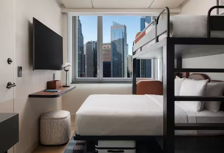 Helles Hotelzimmer mit Doppelbett, Hochbett und Blick aus dem Fenster auf die Skyline.