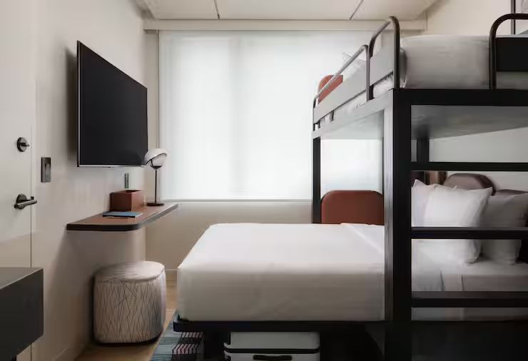 Hotelzimmer mit Doppelbett, Hochbett-Aufsatz, Wandfernseher und Hocker.