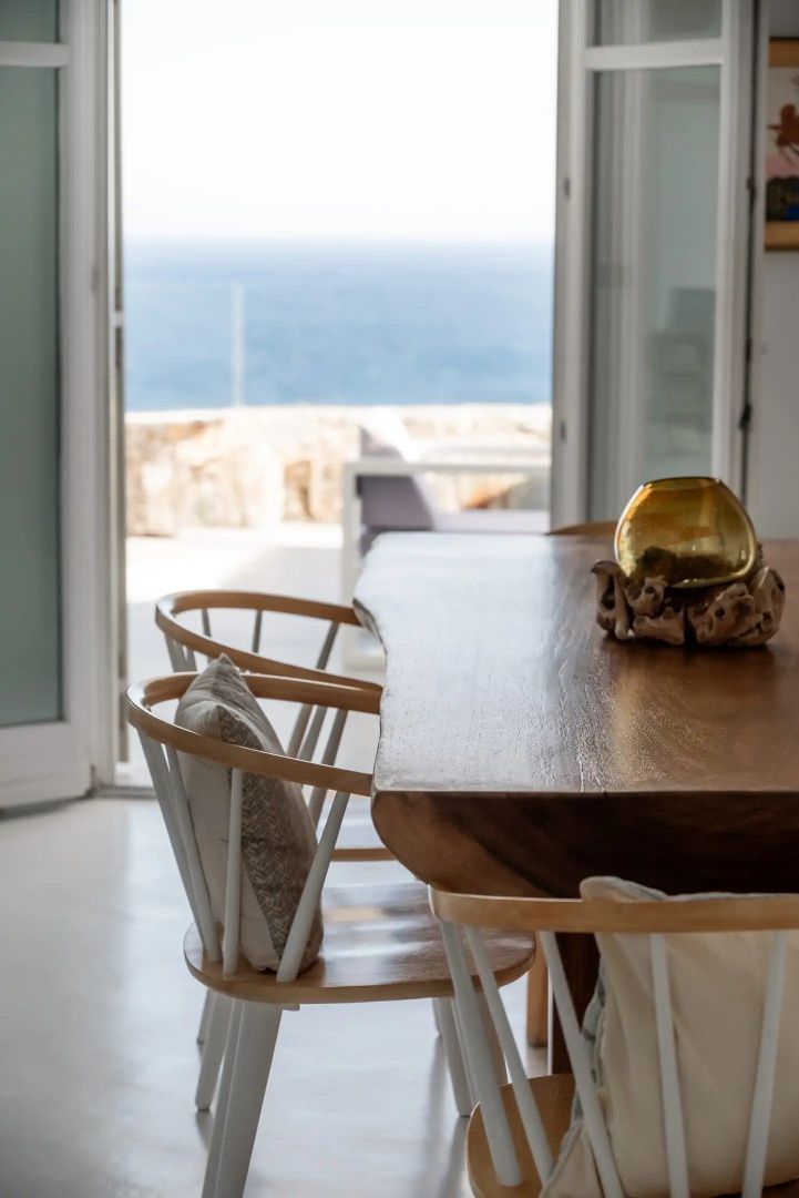 Holztisch mit Stühlen und einer Deko-Vase vor geöffneter Balkontür mit Meerblick.