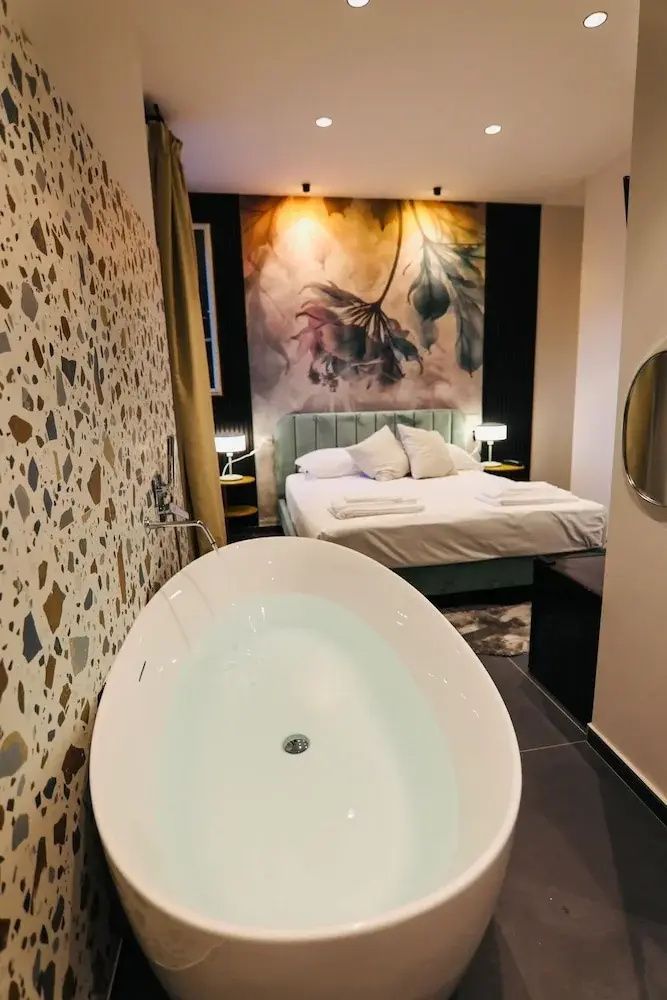 Freistehende Badewanne vor einer bunten Mosaikwand neben einem Doppelbett im Hotelzimmer.