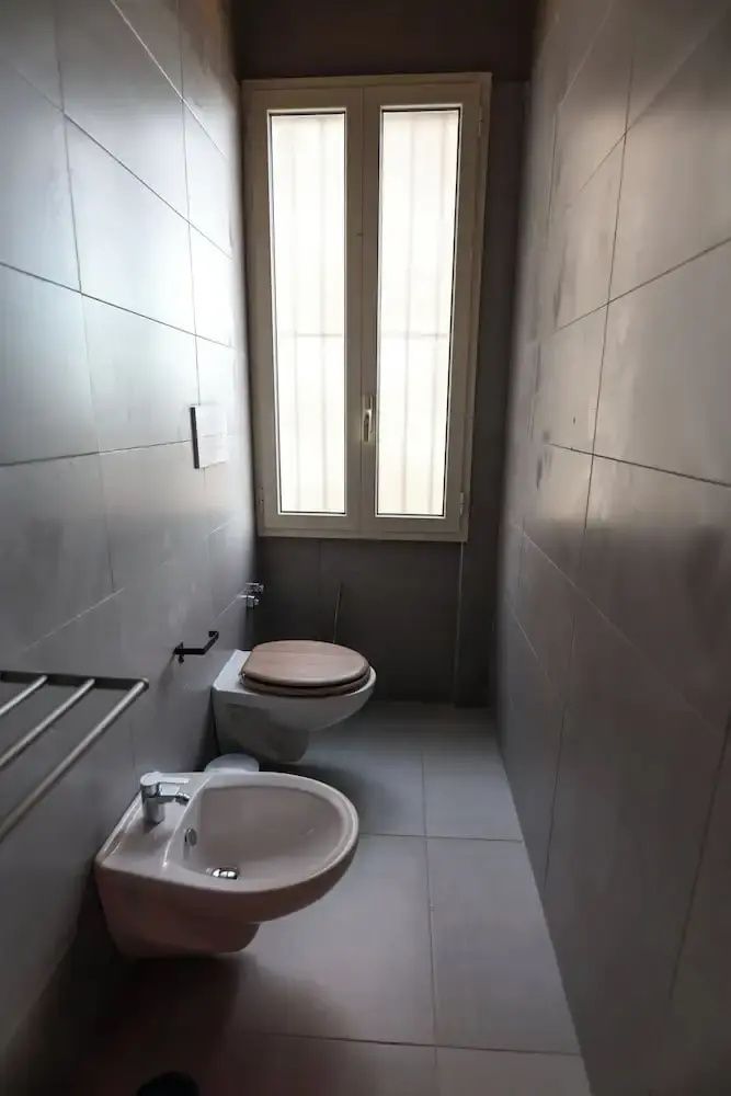 Badezimmer mit Toilette, Bidet, kleinem Waschbecken und heller Fensterfront.
