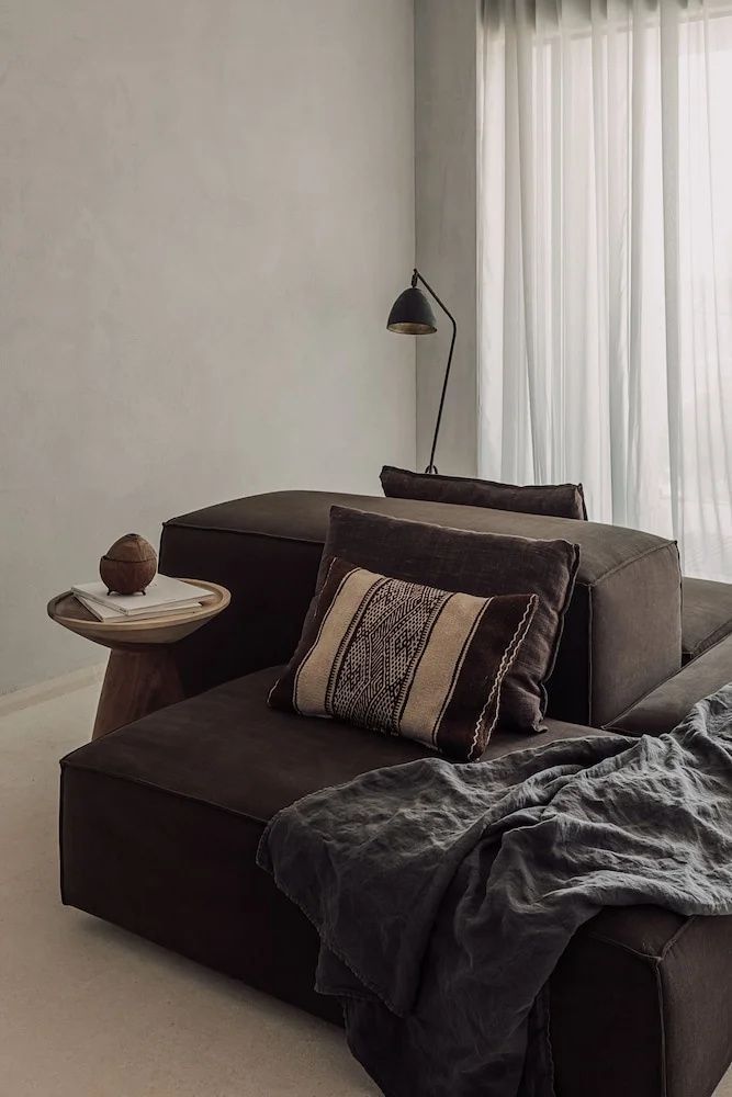Dunkelbraune Couch mit bedrucktem Kissen, Beistelltisch und Stehlampe neben einer Gardine.