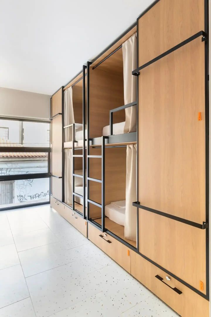 Reihe von Etagenbetten aus Holz mit schwarzen Metallleitern und hellen Vorhängen im Mehrbettzimmer.