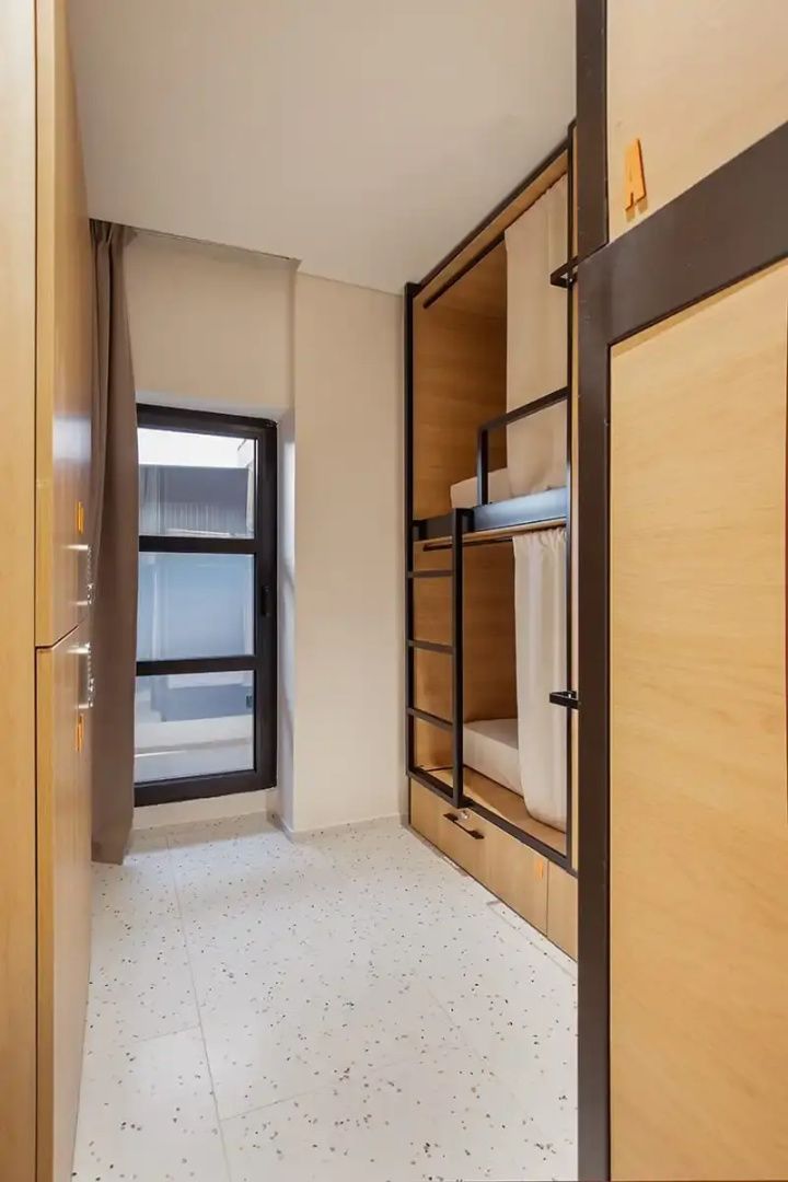 Schmaler Bereich im Schlafsaal mit Etagenbett aus Holz, Schränken und einem hochformatigen Fenster.