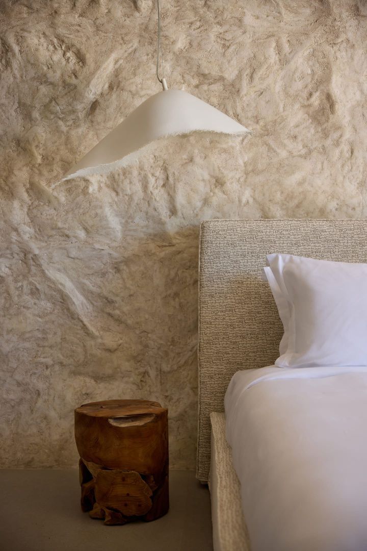 Bett mit hellem Kopfteil und weißer Bettwäsche vor einer strukturierten Wand aus Naturstein.