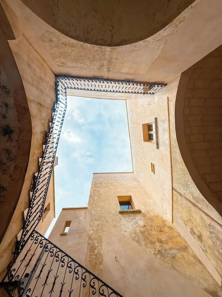 Blick von unten in den Innenhof mit historischen Steinwänden, Treppengeländer und blauem Himmel.