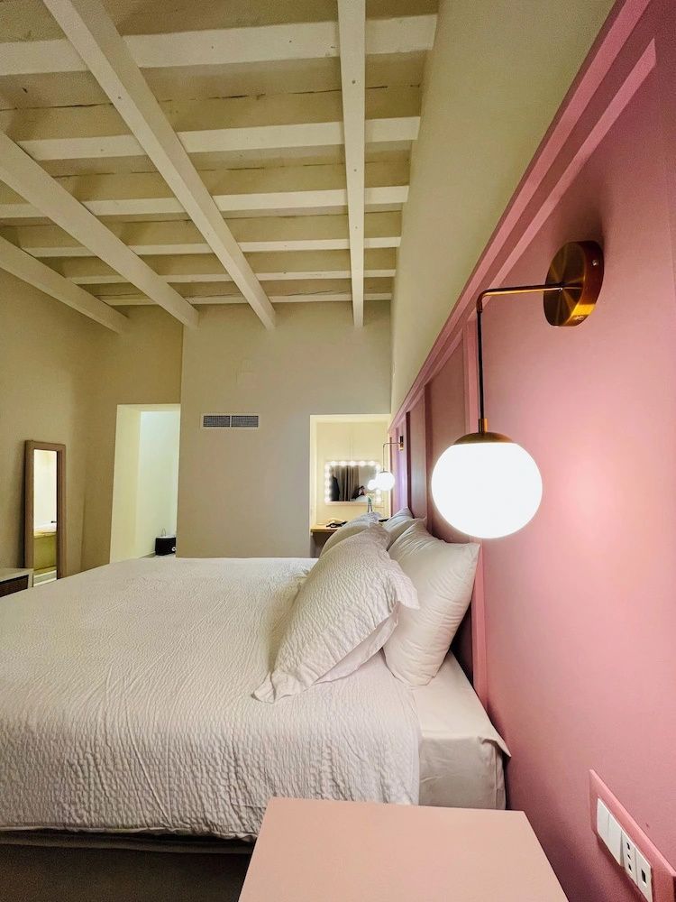 Bett mit weißer Decke vor rosa Wandpaneel und weiße Balkendecke mit Kugelleuchte.