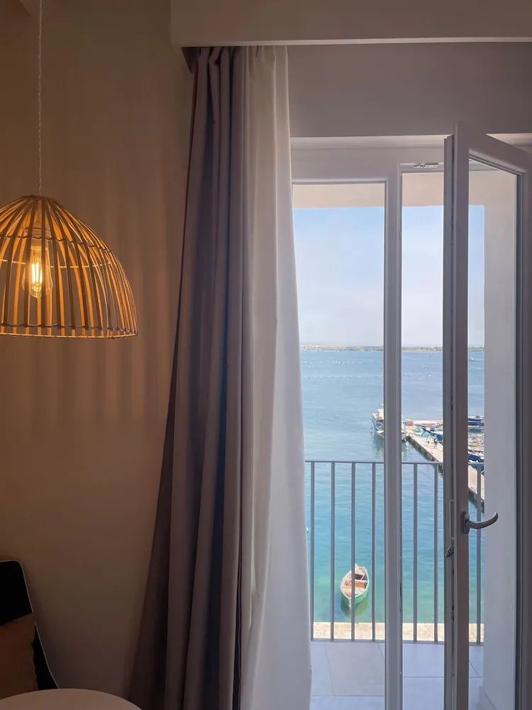 Blick durch ein geöffnetes Fenster auf den blauen Ozean und einen kleinen Hafen.