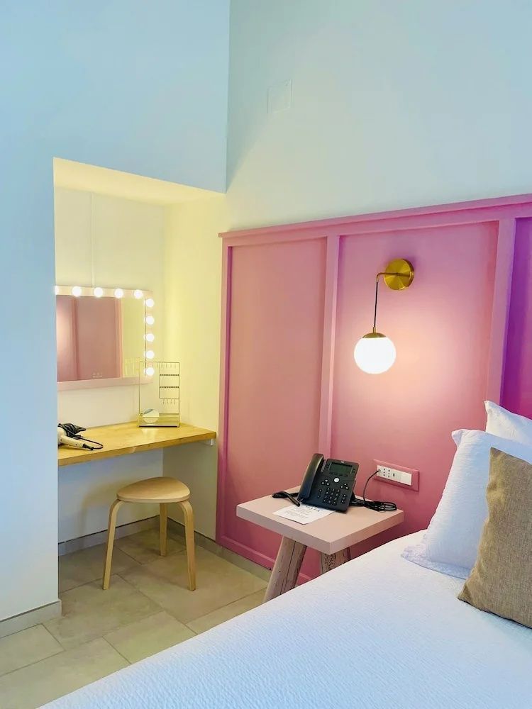 Hotelzimmer mit Bett, rosa Wandoberfläche, Nachttisch und einer kleinen Schminkecke.