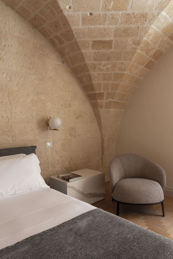 Schlafbereich mit weiß gekleidetem Bett, grauem Sessel und Gewölbedecke aus Naturstein.