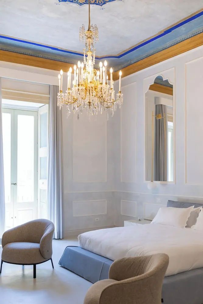 Großes Zimmer mit einem Kronleuchter, verzierter Decke und einem hellen Doppelbett.